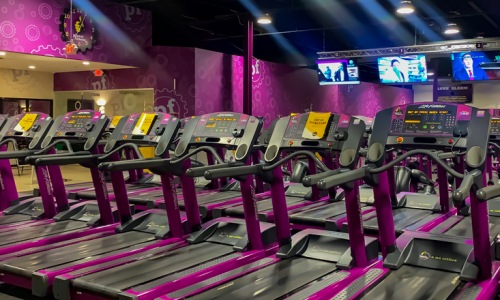 Rows of treadmill exercise machines facing large TV screens at Planet Fitness gym